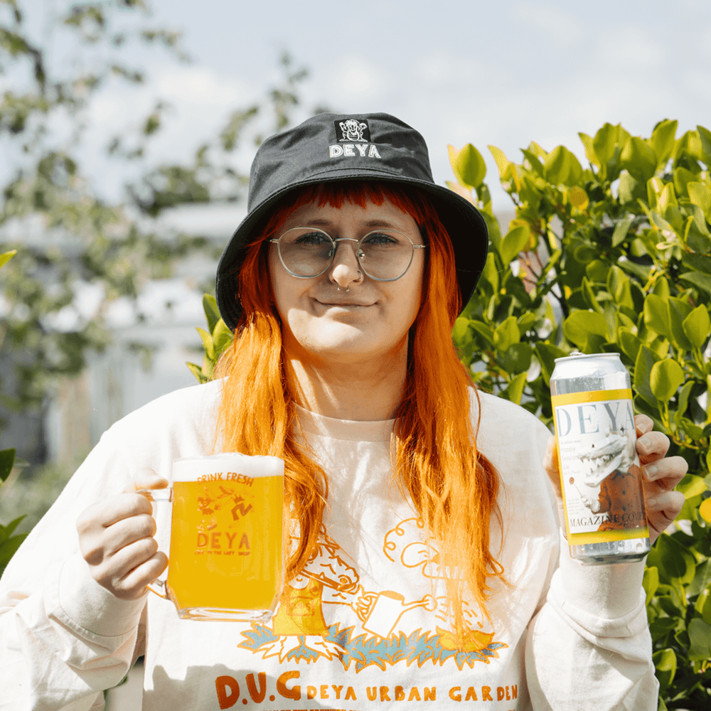 Meg holding a 500ml can of Magazine cover pale ale, a DEYA Drink Fresh Tankard and wearing a Navy "Happy Croc" Bucket Hat
